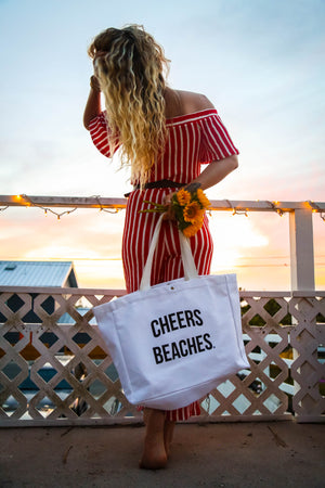 A woman stands on a balcony at sunset wearing a red and white striped off-the-shoulder dress and holding a white canvas tote bag printed with the text 'CHEERS BEACHES.' She is seen from behind, holding sunflowers, with a railing and sky in the background.