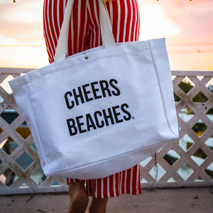 A person wearing a red and white striped dress holds a large white canvas tote bag printed with the words 'CHEERS BEACHES.' in black text, while holding a bouquet of sunflowers on a balcony at sunset.